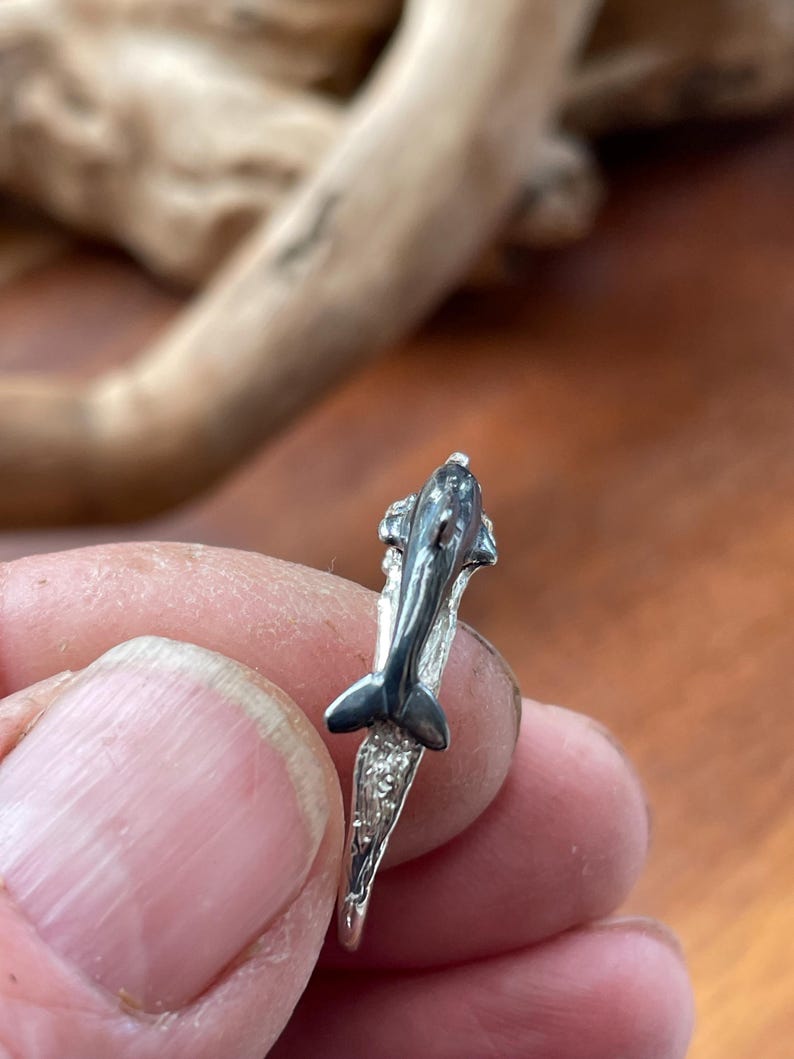 May include: A silver ring featuring a detailed dolphin design. The dolphin is depicted with a sleek, curved body and a prominent dorsal fin. The ring's band has a textured, wave-like pattern, adding to the aquatic theme.