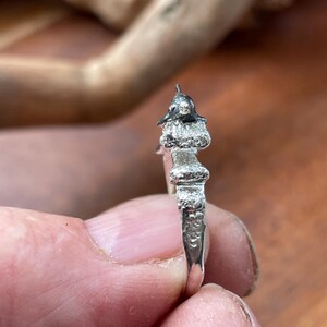 May include: A silver ring with a detailed, miniature sculpture of a figure wearing a hat. The ring's design features textured elements and a polished finish, showcasing intricate craftsmanship. The ring is held between fingers.