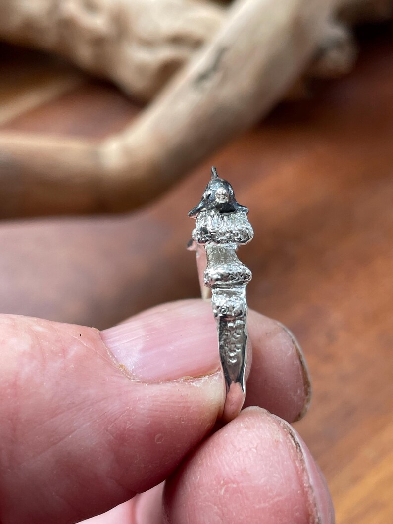 May include: A silver ring with a detailed, miniature sculpture of a figure wearing a hat. The ring's design features textured elements and a polished finish, showcasing intricate craftsmanship. The ring is held between fingers.
