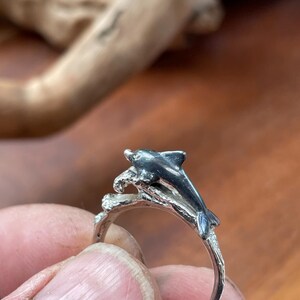 May include: A silver ring featuring a dolphin leaping over a wave. The dolphin is detailed with dark gray accents, and the ring band has a textured, organic design. The ring is held against a blurred wooden background.