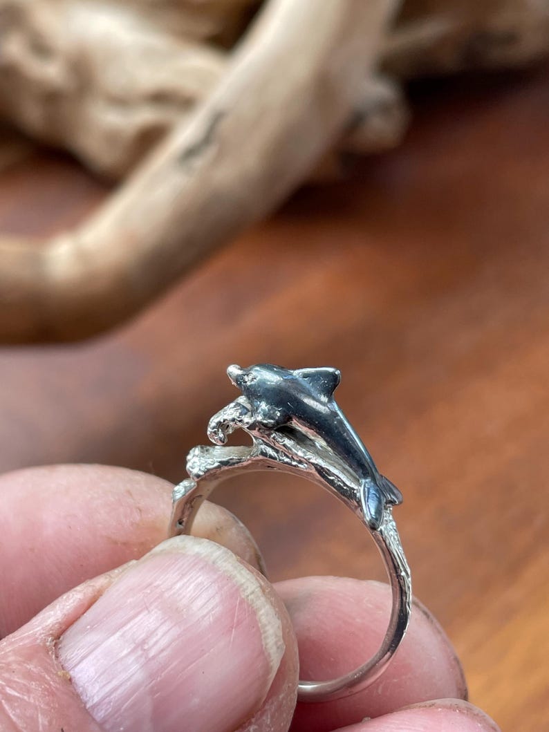 May include: A silver ring featuring a dolphin leaping over a wave. The dolphin is detailed with dark gray accents, and the ring band has a textured, organic design. The ring is held against a blurred wooden background.