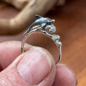 May include: A silver ring featuring a dolphin leaping over a wave. The dolphin has a dark gray finish, and the ring is held in a hand. The ring's design is detailed, with the dolphin and wave clearly defined.