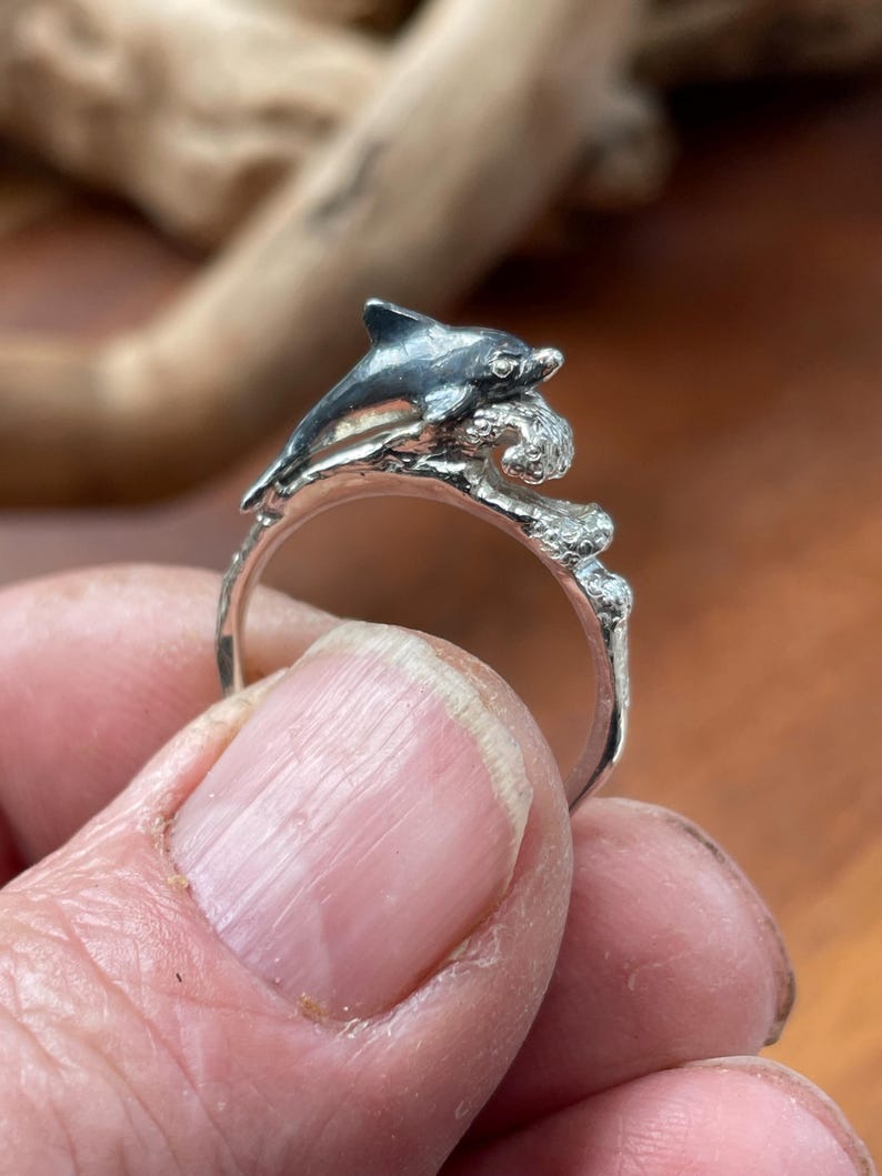 May include: A silver ring featuring a dolphin leaping over a wave. The dolphin has a dark gray finish, and the ring is held in a hand. The ring's design is detailed, with the dolphin and wave clearly defined.