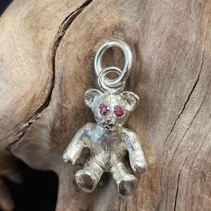 Pode incluir: Um berloque de urso de pelúcia prateado com olhos de pedras preciosas vermelhas. O berloque tem detalhes de patas, orelhas e um laço para prender. O urso tem aproximadamente 2,5 cm de altura.