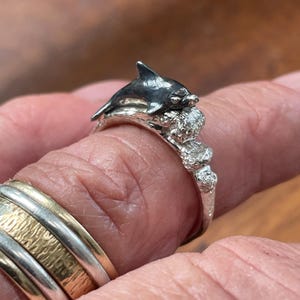 May include: A silver ring featuring a detailed dolphin design. The dolphin is depicted with a dark gray finish, resting on a textured base. The ring's band has a textured, organic appearance, showcasing the craftsmanship.