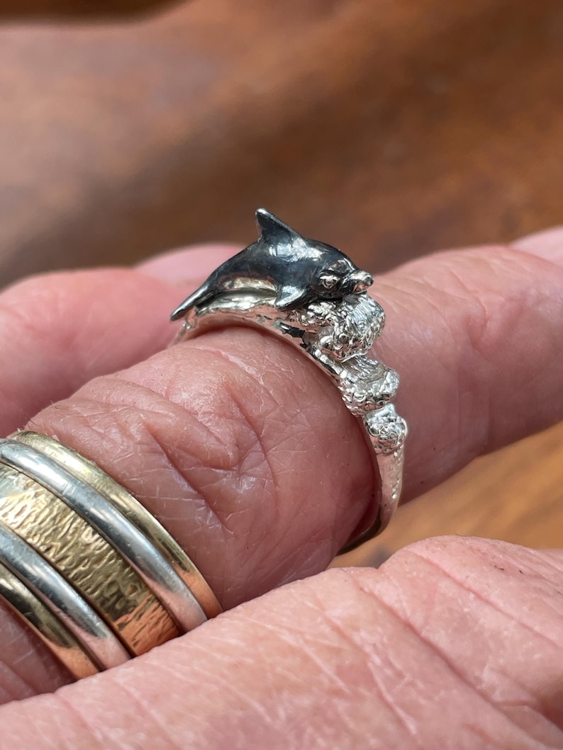 May include: A silver ring featuring a detailed dolphin design. The dolphin is depicted with a dark gray finish, resting on a textured base. The ring's band has a textured, organic appearance, showcasing the craftsmanship.