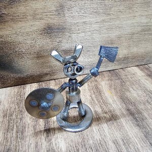 May include: A small metal figurine constructed from hardware components including nuts, bolts, and washers. The figure features a wing nut head, a threaded bolt torso, and legs made of bolts. It holds a small metal axe and a circular shield.