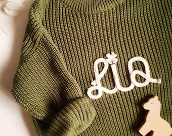 May include: Olive green knit sweater with the name "Lia" in white rope lettering. A small wooden horse figurine rests beside the sweater. The sweater has a ribbed texture and a crew neck.
