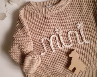 May include: A beige knitted sweater with the word "mini" embroidered in white yarn. A small wooden toy horse is on the sweater.