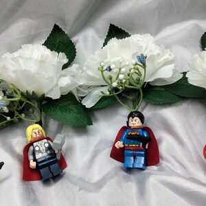 May include: Four miniature superhero figurines, including Hulk, Thor, Superman, and Captain America, are attached to white carnation buttonholes.