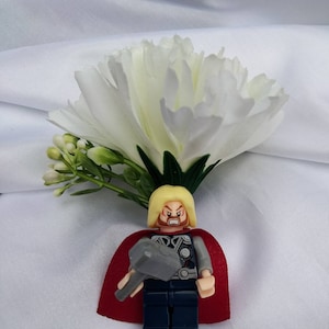 May include: A Lego figurine of Thor, wearing a red cape and holding a hammer, is placed on top of a white carnation.