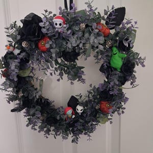 May include: A circular wreath featuring dark grey eucalyptus foliage, black fabric roses, and small orange pumpkins. The wreath is decorated with miniature character figures including a skeleton in a Santa hat, a green ghost, and a couple figure.