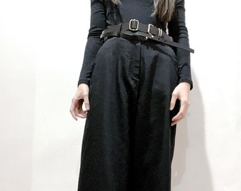 Könnte beinhalten: Ein schwarzes Langarmshirt mit einem schwarzen Ledergürtel mit silbernen Schnallen. Die Person trägt eine schwarze, weite Hose und schwarze Stiefel.