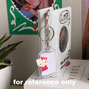 May include: A white binder with a clear plastic photo holder featuring a photo of a person with the word "CUTE" written above it. The binder has a silver keychain with a clear plastic photo holder featuring a Hello Kitty character. The keychain has a silver ring with the word "COLLECTOR'S" written on it. The binder has the text "Why did you show up now?" and "HOW SELF-LOVE IS VIBES?" written on it.