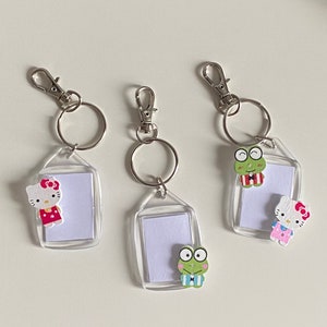 May include: Three silver keychains with clear plastic frames and colorful cartoon character charms. Each keychain has a small, clear plastic rectangle for inserting a photo or note. The charms include a green frog, a pink and white Hello Kitty, and a pink and white Hello Kitty.