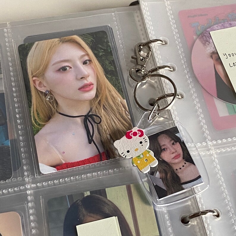 May include: A clear plastic binder with a silver ring mechanism. A photo card of a woman with blonde hair is in one of the pockets. A keychain with a Hello Kitty charm is attached to the binder.