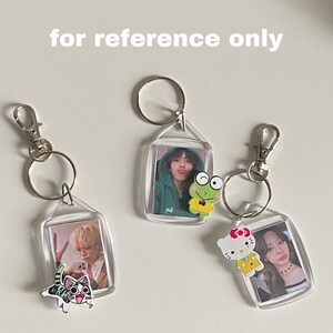 May include: Three clear plastic keychains with photos inside. Each keychain has a small wooden charm attached: a frog, a cat, and Hello Kitty. The keychains are on a white surface.