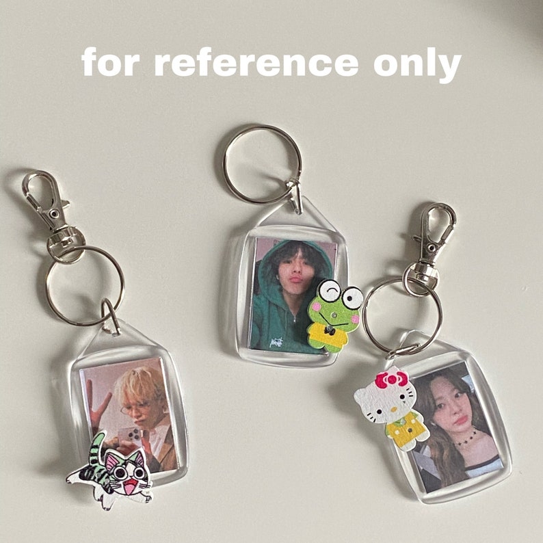 May include: Three clear plastic keychains with photos inside. Each keychain has a small wooden charm attached: a frog, a cat, and Hello Kitty. The keychains are on a white surface.