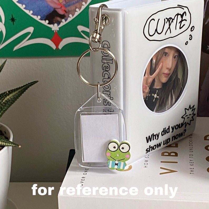 May include: A white photo album with a clear plastic keychain attached. The keychain has a small frog charm and a blank photo card. The album has the text "Collector's" printed on the side and "Cutie" written in black marker on the front. The album also has the text "Why did you show up now?" and "How's" printed on the front.