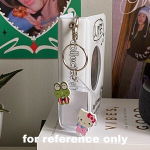 May include: White photo album with a clear plastic keychain attached. The keychain has a small frog and Hello Kitty charm. The album has the word "CUTE" written on it.