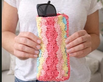 May include: A handmade, colorful crochet sunglasses case. The case is a rectangular shape with a textured pattern in shades of pink, yellow, blue, and cream. Black sunglasses are visible inside the case. The case is held by a person.