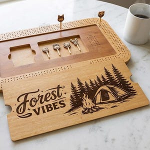 K&ouml;nnte beinhalten: Cribbage-Brett aus Holz mit tierf&ouml;rmigen Stiften und einem dekorativen Deckel mit einer Waldszene und dem Text "Forest Vibes". Enth&auml;lt W&uuml;rfel und eine Spielkarte. Das Brett ist ca. 30 cm lang.