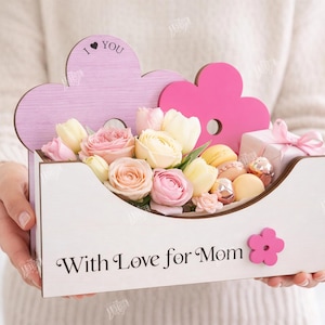 May include: A wooden flower box with the text "With Love for Mom" and a pink flower accent. The box is filled with pink and yellow flowers, macarons, and a small gift box. The top of the box has a pink cloud-shaped design with the text "I &hearts; YOU".
