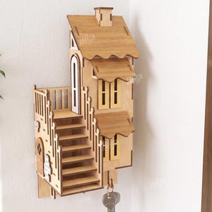 May include: A light brown wooden wall-mounted key holder shaped like a multi-story house with a staircase, balcony, and windows. The design includes text stating CUT READY FILE DIGITAL DOWNLOAD and MATERIA WIDTH: 3MM, with a metal key hanging below.