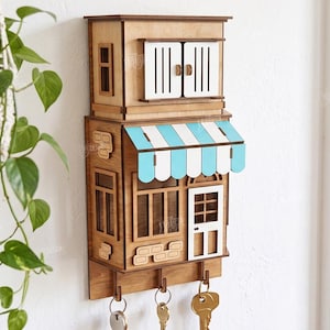 May include: A wall-mounted wooden key holder shaped like a two-story building with a light blue and white striped awning, white window shutters, a white door, and four metal hooks at the bottom for hanging keys, designed for 3mm material thickness.