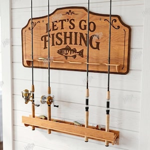 May include: Wooden fishing rod holder with the text "LET'S GO FISHING" and a fish graphic. The holder has slots for rods and a lower shelf for lures. Several fishing rods are displayed. The sign is mounted on a white wall.