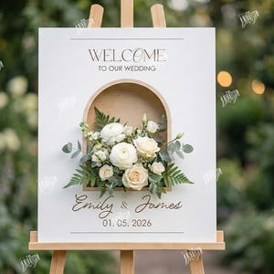 May include: A white wedding welcome sign on an easel, featuring a floral arrangement in a wooden arch. The sign reads "Welcome to our Wedding" and includes the names "Emily & James" and the date "01.05.2026".