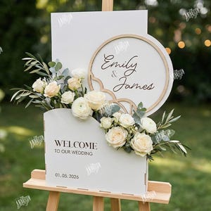 May include: A white wedding sign on a wooden easel, featuring the names "Emily & James" and the date "01.05.2026". The sign is decorated with flowers and greenery, with a wooden frame and a scalloped edge.