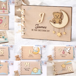 May include: Wooden baby memory books with laser-cut designs. One book features a bear on a crescent moon, stars, and the name "James." Other books have names like "Amelia," "Emily," and "David." The books are tied with ribbons.