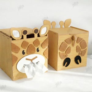 May include: Two wooden tissue box holders shaped like giraffes. One has tissues coming out of the mouth. The giraffe faces are made of light and dark brown wood. The boxes are on a white surface. The text on the right says "CUT READY FILE".