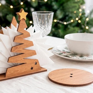 May include: A wooden napkin holder shaped like a stylized Christmas tree with a gold star topper and two small star cutouts at the base, holding white napkins, accompanied by a circular wooden coaster with two small star cutouts, 3mm material width.