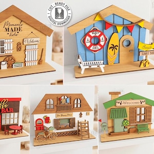 Puede incluir: Una colección de cinco diseños de casas de madera cortadas con láser. Los diseños incluyen una casa con el texto "Moments Made Here", una casa con temática de playa, una cafetería, una casa con una bicicleta y una casa con un letrero de bienvenida.
