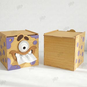 May include: Two wooden tissue box holders. One is decorated with a cartoon monster face with a single eye, teeth, and purple accents. The other is plain. The boxes are made of light brown wood.