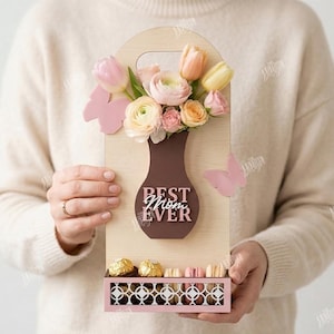 May include: A wooden gift box with a vase-shaped cutout holding flowers and the text "BEST MOM EVER." The box also features a lower compartment with chocolates. Pink butterflies and flowers add a decorative touch.