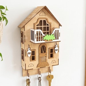 May include: A light-colored wooden wall-mounted key holder shaped like a house with a balcony, a central door, and two white lantern-style accents. The structure features three metal hooks at the bottom holding keys and a 3mm material width label.