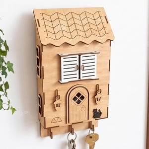May include: A light-colored wooden wall-mounted key holder shaped like a house with a tiled roof, white shutters, a central door, two hanging lanterns, and a small black cat silhouette. Three metal hooks at the bottom hold several silver keys.