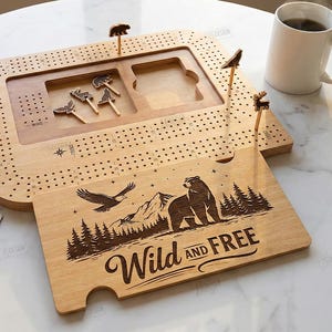 K&ouml;nnte beinhalten: Ein h&ouml;lzernes Cribbage-Brett mit tierf&ouml;rmigen Stiften und einem separaten Holzst&uuml;ck mit den Worten "Wild and Free" und einer B&auml;ren- und Adlerillustration. Enth&auml;lt W&uuml;rfel, Spielkarten und eine Kaffeetasse.