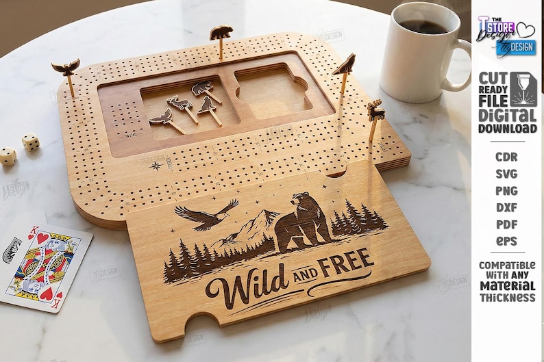 K&ouml;nnte beinhalten: Ein h&ouml;lzernes Cribbage-Brett mit tierf&ouml;rmigen Stiften und einem separaten Holzst&uuml;ck mit den Worten "Wild and Free" und einer B&auml;ren- und Adlerillustration. Enth&auml;lt W&uuml;rfel, Spielkarten und eine Kaffeetasse.