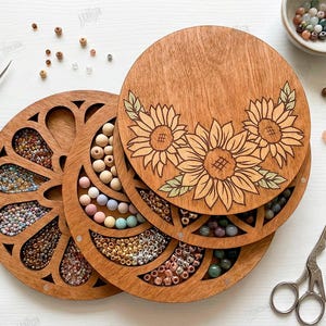 May include: Wooden bead organizer with sunflower design. The organizer has multiple compartments filled with colorful beads. A pair of tweezers and small scissors are also present. The organizer is made of wood and has a natural finish.