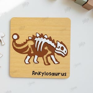 K&ouml;nnte beinhalten: Holzpuzzle mit einem braunen Ankylosaurus-Dinosaurier mit wei&szlig;en Knochen-Details. Das Puzzle befindet sich auf einem hellen Holzmaserungshintergrund. Der Text "Ankylosaurus" steht unten. Eine Hand h&auml;lt Puzzleteile.