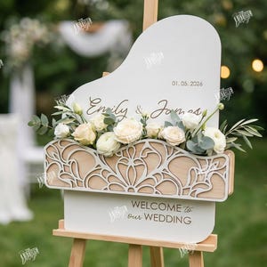 May include: A wooden wedding sign with a white top and a decorative floral arrangement. The sign reads "Emily & James" and "Welcome to Our Wedding". The date "01.05.2026" is also displayed. The sign is supported by a wooden easel.
