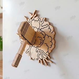Puede incluir: Una decoraci&oacute;n de pared de madera clara que muestra un martillo incrustado en un patr&oacute;n circular en forma de nube con contornos negros grabados. La pieza est&aacute; dise&ntilde;ada para un grosor de material de 3 mm e incluye archivos digitales.