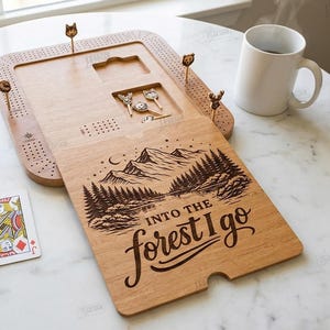 K&ouml;nnte beinhalten: Holz-Cribbage-Brett mit eingravierter Berg- und Waldszene, W&uuml;rfeln, Spielkarten und dekorativen Stiften. Der Text "Into the forest I go" ist auf dem Brett zu sehen. Eine wei&szlig;e Tasse ist ebenfalls vorhanden.