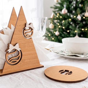 May include: A triangular wooden napkin holder featuring two cutout Christmas ornament designs with bows, holding white napkins. Beside it sits a circular wooden coaster with a matching ornament cutout. Text on the right indicates 3MM material width.
