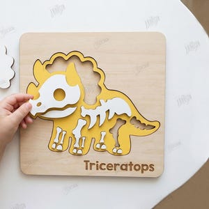 K&ouml;nnte beinhalten: Ein h&ouml;lzernes Triceratops-Puzzle mit gelbem und wei&szlig;em Dinosaurierskelett-Design. Das Puzzle befindet sich auf einer hellen Holzbasis, mit dem Wort "Triceratops" unten. Das Bild zeigt auch die Hand einer Person, die ein Puzzleteil einsetzt.