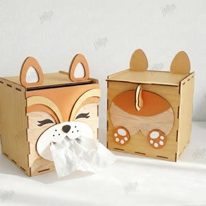May include: Two wooden tissue box holders, one with a fox face design and the other with a fox tail and paw design. The boxes are made of light brown wood with orange and white accents. The text on the side says "CUT READY FILE" and "MATERIA WIDTH: 3MM".